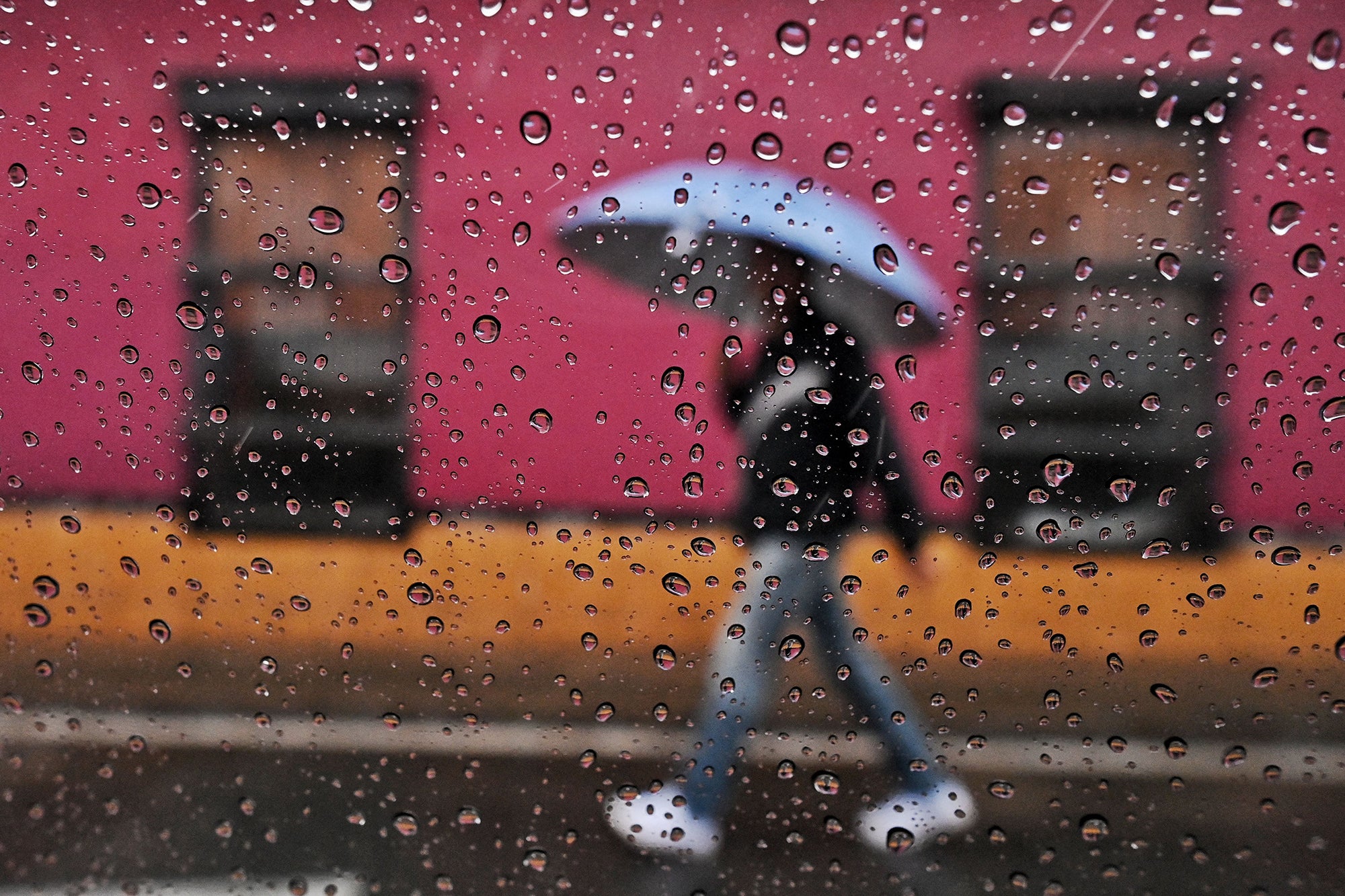 A photograph focused on water droplets on a window, blurry in the background, a man walks holding an umbrella under the rain in front of a pink and yellow wall