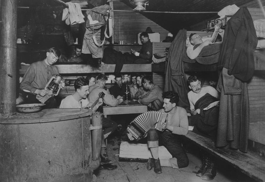 A collection of men relaxing in what looks to be a sort of cabin. Several play cards, one plays an accordion.