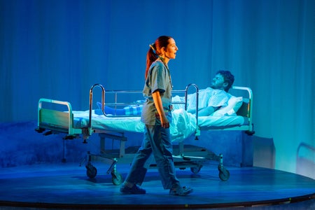 On stage, a woman in scrubs walks past a man in a hospital bed