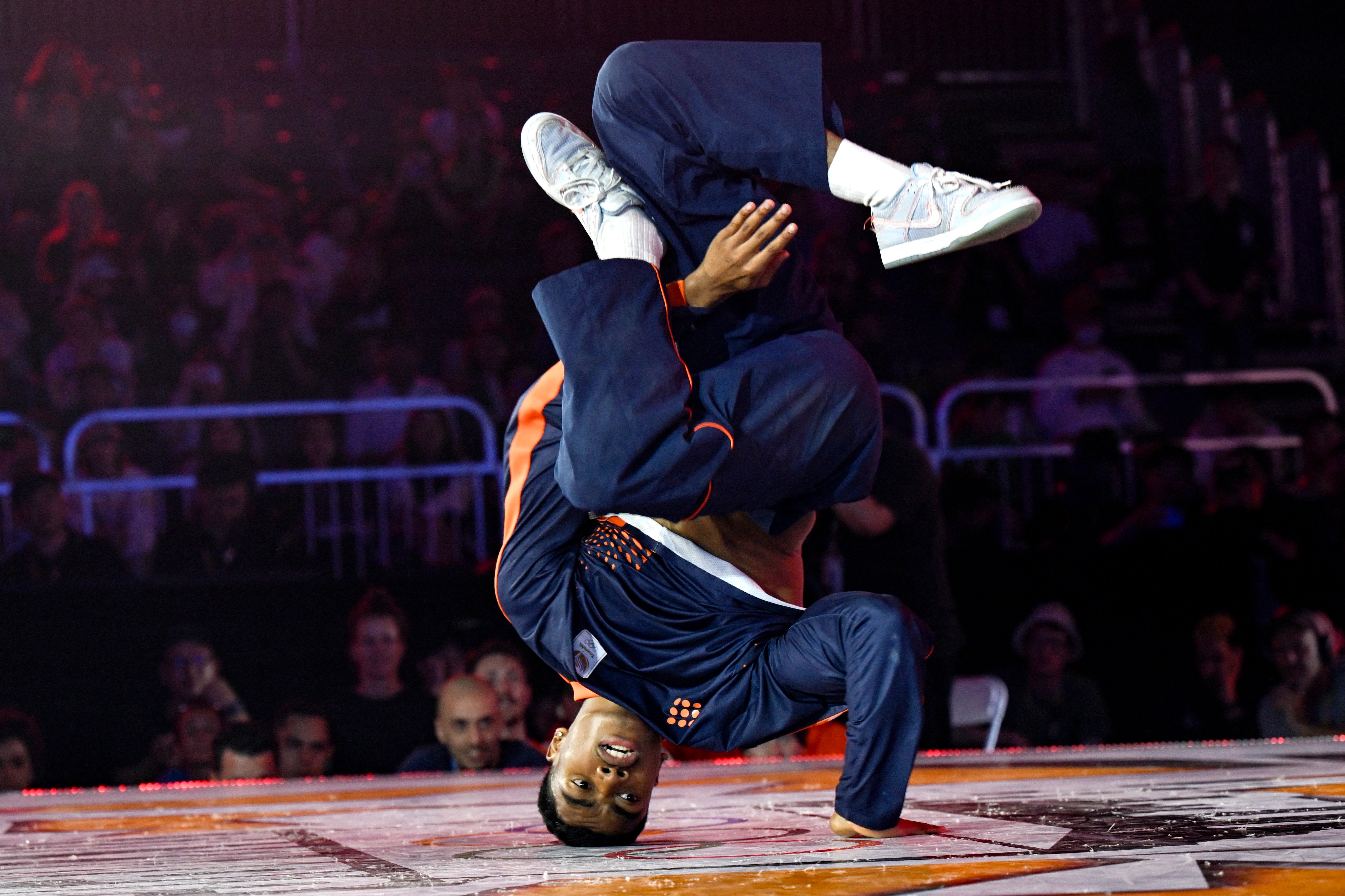 Breakdancer on his head with twisted arms and legs in blue sports suit with orange trim.