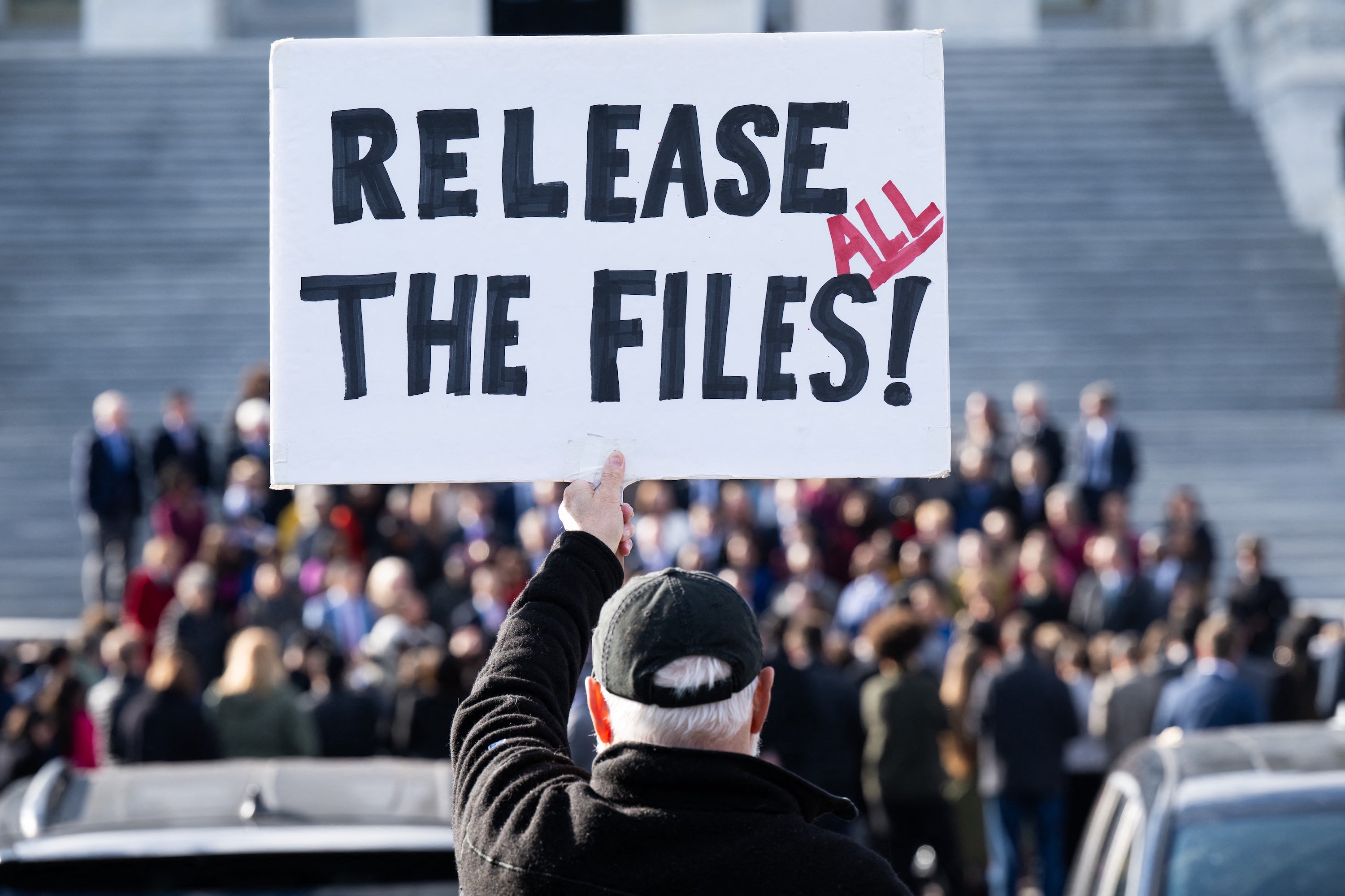 A white sign is held by a man with his back to viewer in from of capitol steps crowd.