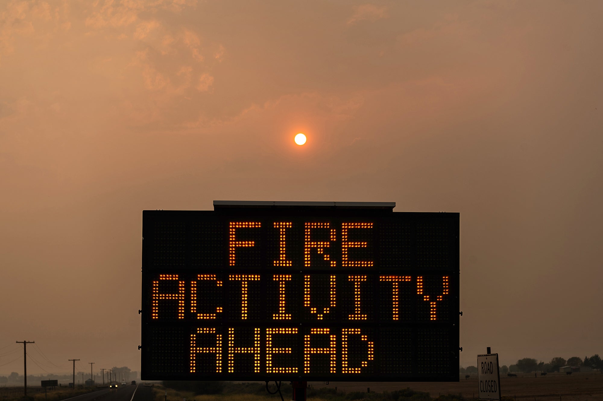 A highway sign reads, "FIRE ACTIVITY AHEAD," in the foreground, the landscape, sky, and sun in the background can be seen shrouded with heavy wildfire smoke, causing a warm, orange tint to be cast