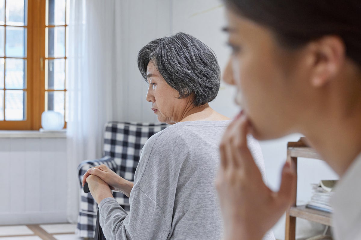 Older adult parent appears in distress sitting with their back towards the camera in the middle of the frame, to the right, in the foreground and out of focus, a younger adult in profile touches their mouth