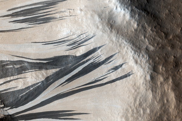 Many dark and light-toned slope streaks are visible on the wall of the trough surrounded by dunes in an area of Mars called Acheron Fossae