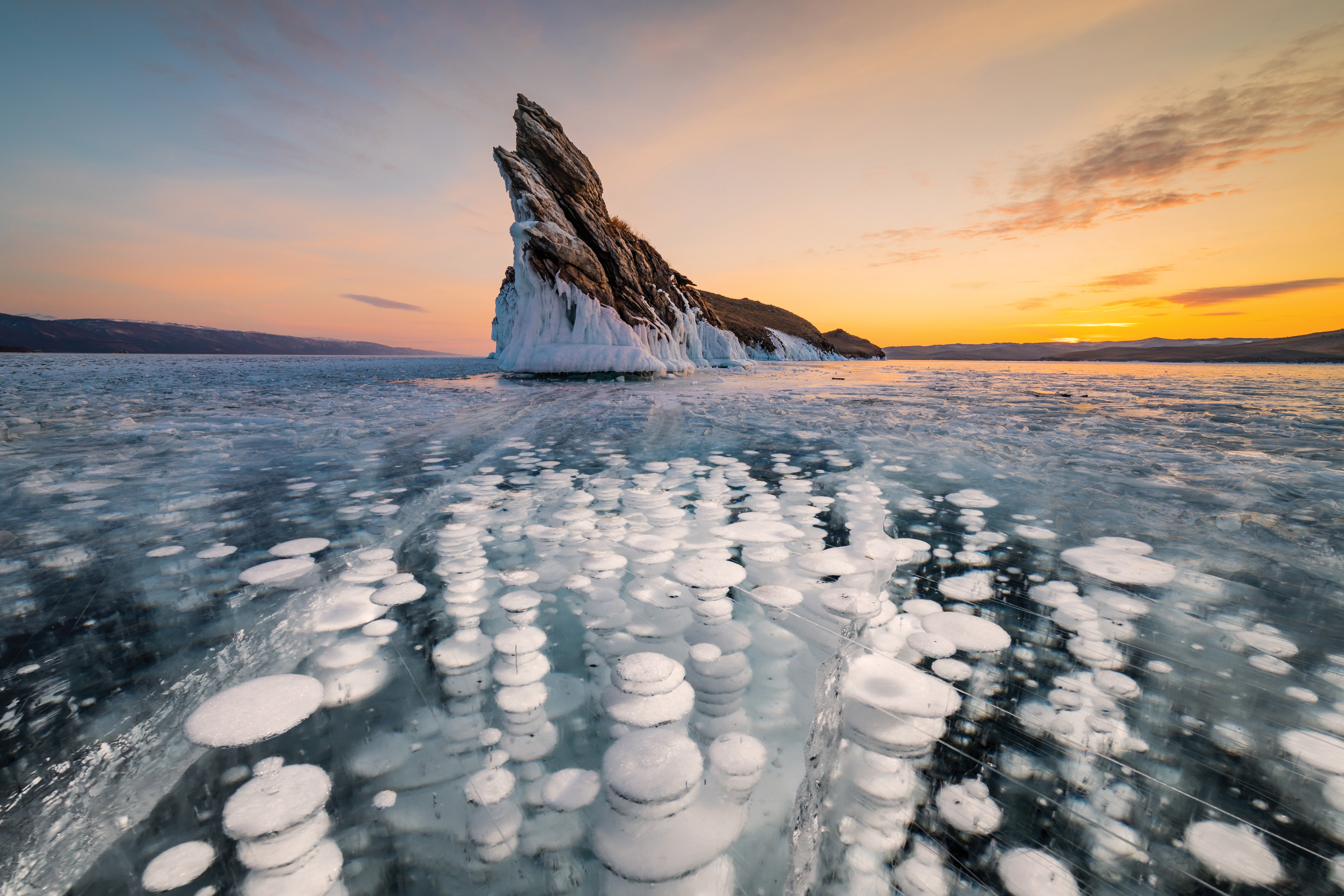 Image of ice bubbles beneath a body of water, with a cliff in the background with a sunset