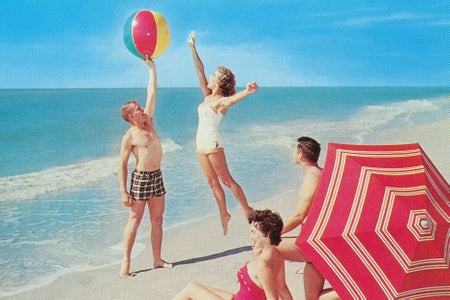 A couple sits under an umbrella at the beach watching another couple play with a beach ball