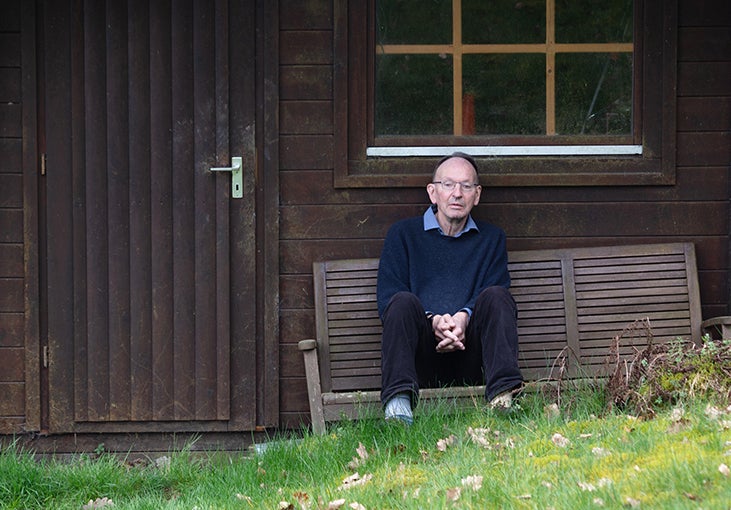 A man sitting with hands folded on a bench outside a wooden shed.