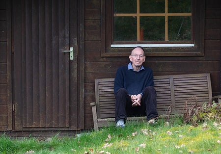 A man sitting with hands folded on a bench outside a wooden shed.