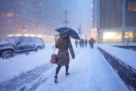 A person in puffer coat and holding an umbrella walks down a snow-covered city sidewalk as snow flies around