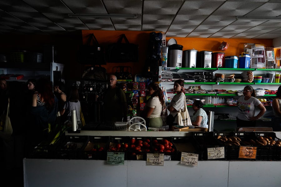 People wait in line to shop for groceries in a dark Lisbon shop without electricity lit only by the light entering from a window out of view to the right of the frame during a widespread power outage that struck Spain and Portugal