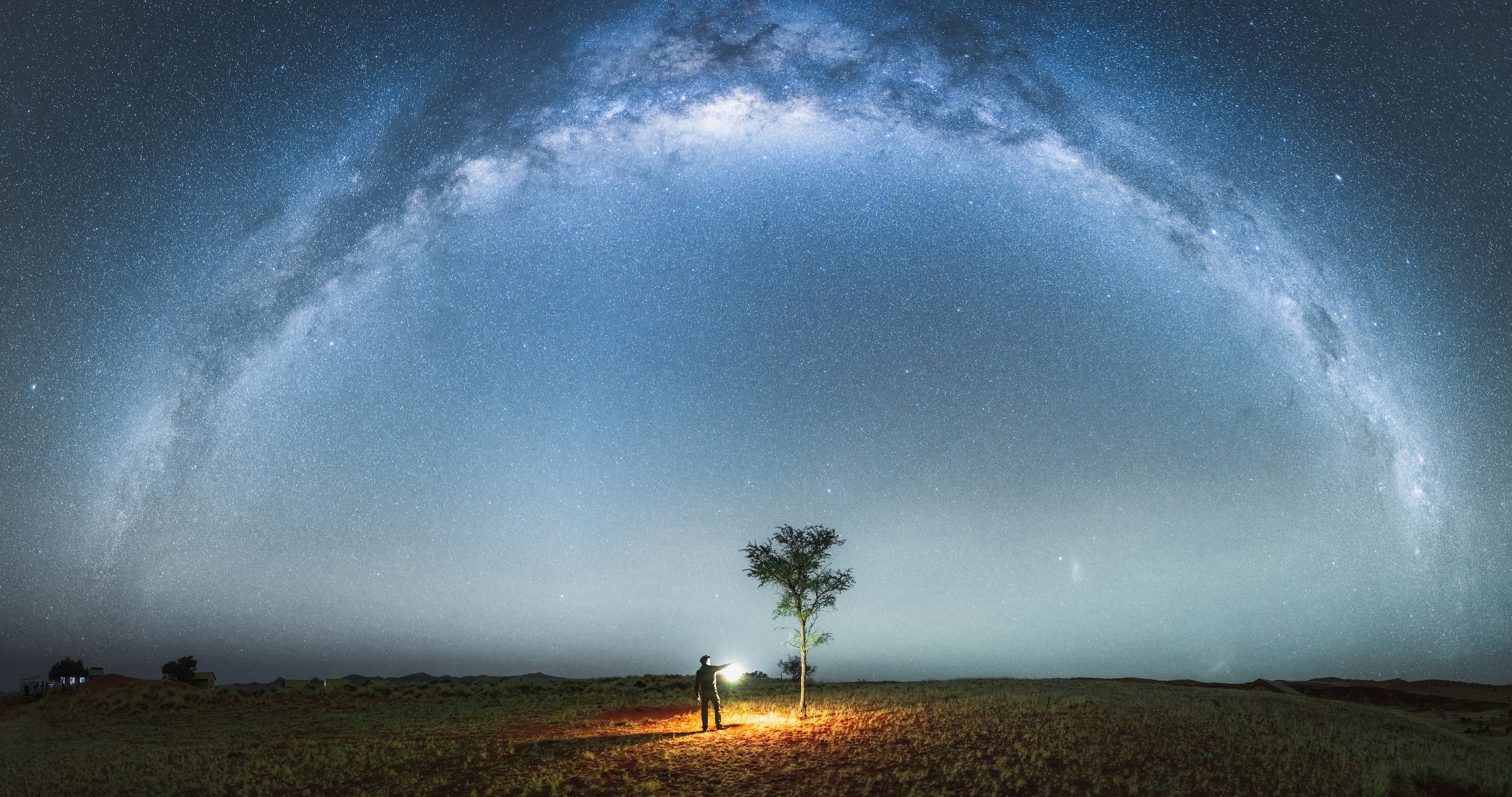 A human figure holds a lantern in a dark, panoramic landscape beneath a starry sky dominated by the arc of the Milky Way
