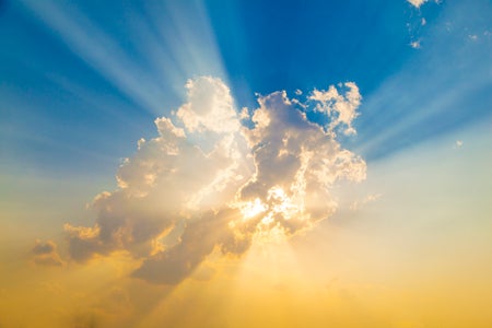 A view of the sun behind a cloud with rays of light , shown against a blue and yellow sky.