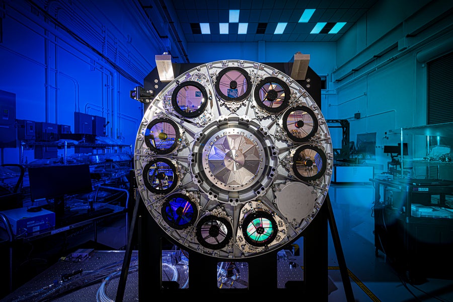A colorful image of the Nancy Grace Roman Space Telescope's element assembly wheel. It appears like a silvery metal wheel, with a star-like conical center surrounded by ten small, black-rimmed circles and one blank circle in the middle. Each of the elements reflects light in shades of blue and turquoise. The element assembly wheel is supported on a frame against the background of a dark clean room, dimly lit by deep blue and turquoise light. Frames, monitors, machinery, and equipment are visible in the background behind the instrument.