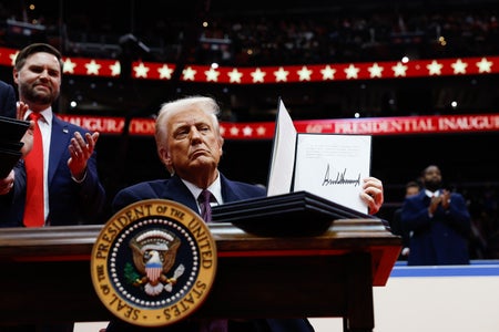 U.S. President Donald Trump sitting at a desk with the presidential seal, holds up an executive order after signing it, with Vice President JD Vance standing behind him, during an indoor inauguration parade at Capital One Arena on January 20, 2025 in Washington, DC.