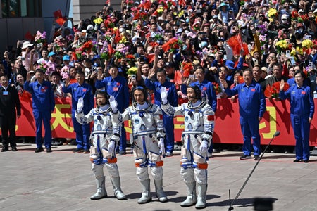 Three Chinese astronauts wave in front of a crowd of public onlookers and space agency officials.