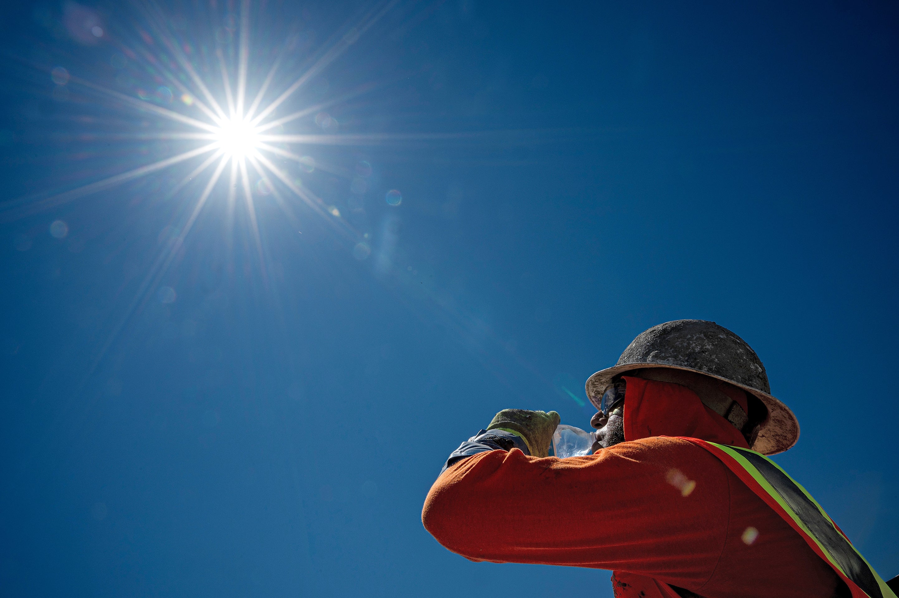 View from below of a construction worker drinking from a water bottle against a blue sky and glaring sun