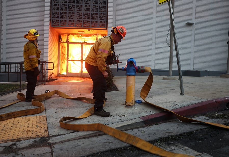 Los Angeles County firefighters try unsuccessfully to get water from a hydrant as they battle the Eaton Fire on January 08, 2025 in Altadena, California. Fire can be seen filling the double glass doorway in the entrance of the building behind the firefighters