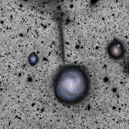 A spiral galaxy seen in full color against a black-speckled grey background. A black stream extends nearly straight up from the galaxy.