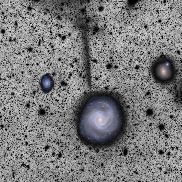 A spiral galaxy seen in full color against a black-speckled grey background. A black stream extends nearly straight up from the galaxy.