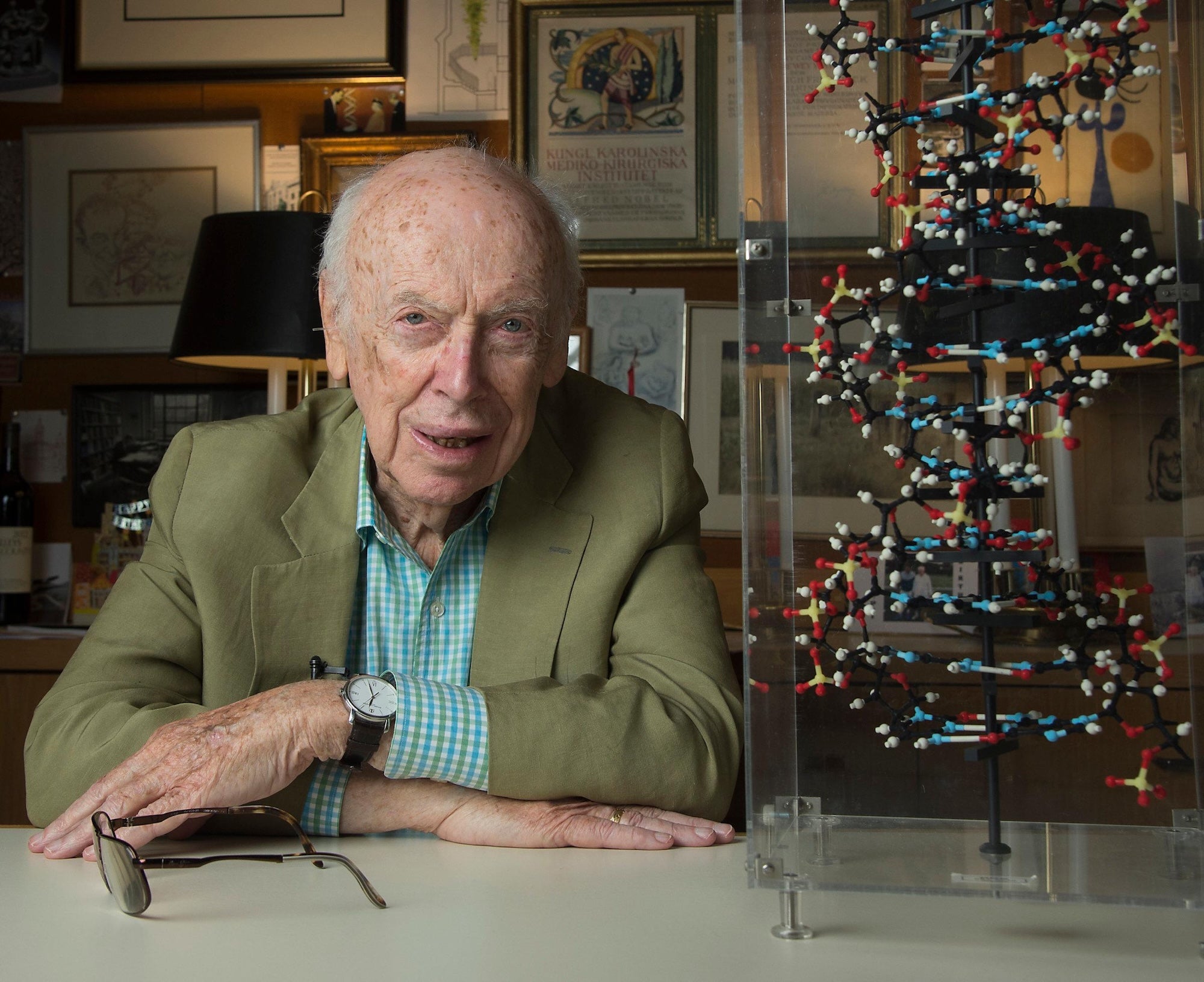 James Watson sitting at a desk on the left, seen from the chest up, his ams folded on the desk. Next to him is a model of a DNA double helix