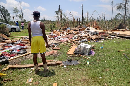 Person in yellow shorts and white tank top with cap looking at a destroyed home.