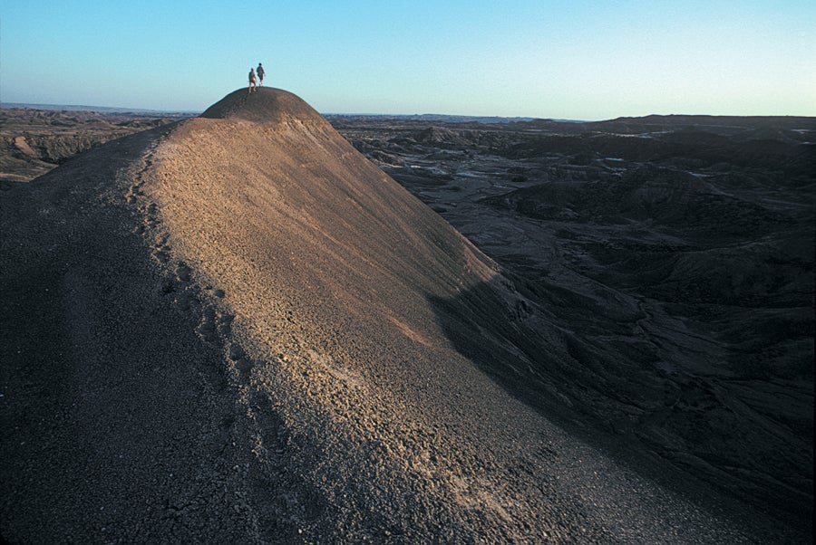 A rugged desert landscape, with two people walking on the top of a sandy hill