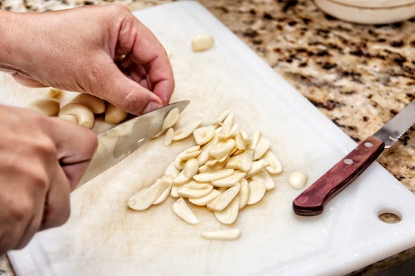 Point of view of female hand cutting garlic on thin slices on plastic cutting board with the aid of a large knife