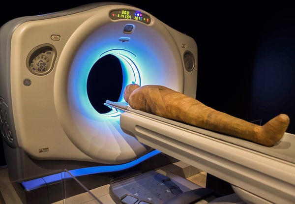 A CT scanning machine in a dark room with a blue light glowing around the center opening of the machine as a linen wrapped mummy lays on the scanner's positioning board