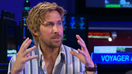 Ryan Gosling wears a blue striped sweater and holds his hands up in front of his face as he explains something. He sits in front of the Mission Control dashboard at NASA's Jet Propulsion Laboratory.