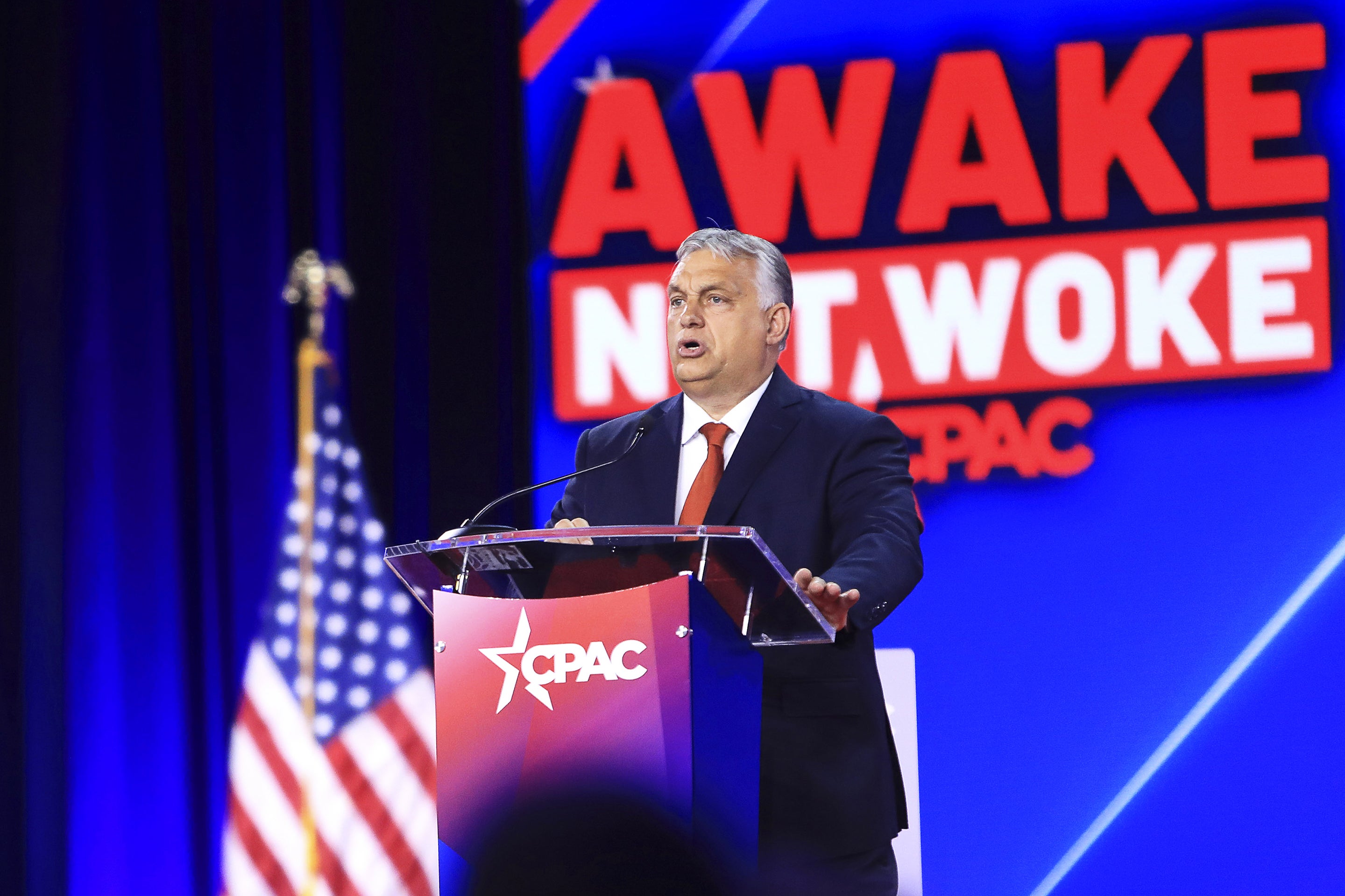 Viktor Orban speaking and standing at a podium on the stage at CPAC in Dallas, Texas in 2022. To the left in the background is an American flag. Directly behind Orban is a large screen displaying text that reads, "AWAKE NOT WOKE," with CPAC's logo beneath it