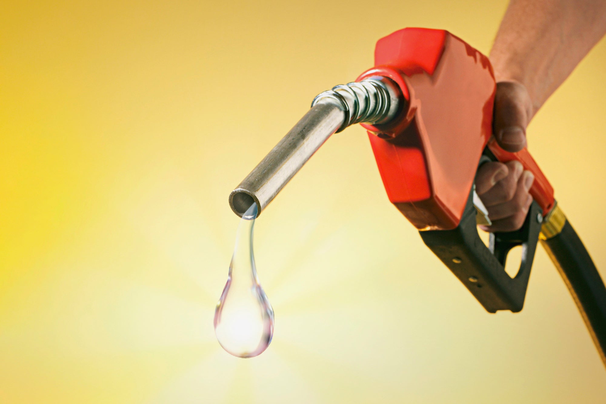 A still-life photograph showing a hand holding a gas pump with a perfectly formed drip of as emerging from the pump nozzle.