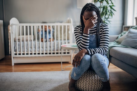 Exhausted mother sitting at home, her crying baby boy is in a crib behind her