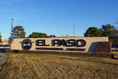 Large stone and stucco entrance sign reading “El Paso International Airport” with a circular airplane logo, set against a clear blue sky with trees in the background and a stop sign visible to the left.