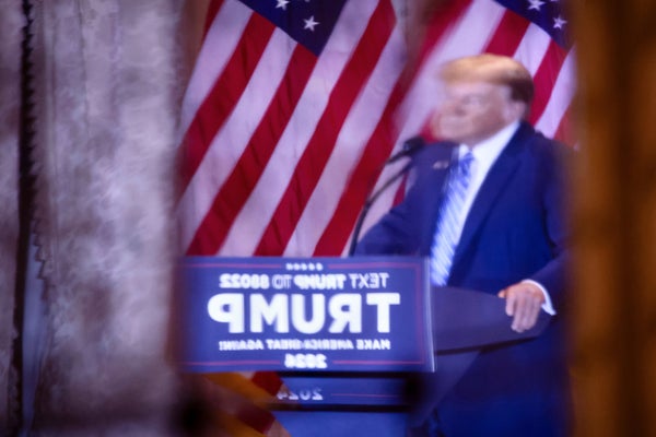 Photograph of a reflection of Donald Trump on stage during a campaign watch party, he is standing at a podium with a sign that reads in all capitalized letters, "TRUMP," behind him are American flags. The reflection has blurred and distorted the image