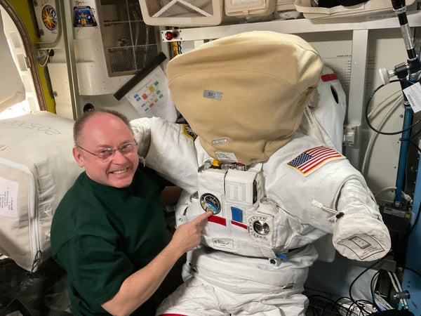 NASA astronaut Mike Fincke poses inside the International Space Station’s Quest airlock next to a spacesuit. The helmet is secured with a protective cover designed to prevent scratches and contamination when the suit is not in use, ensuring the visor remains clear for spacewalks.