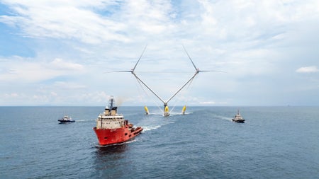 A red boat pulls an offshore wind turbine with two sets of blades anchored to a central platform over the ocean