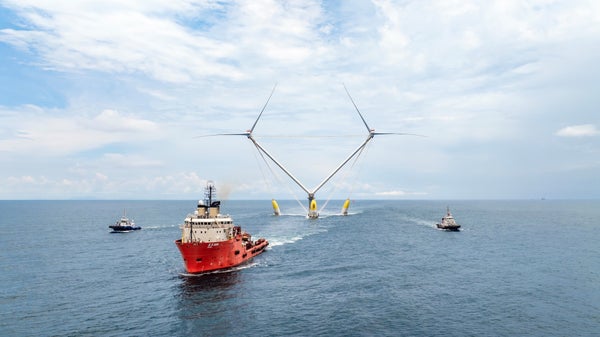 A red boat pulls an offshore wind turbine with two sets of blades anchored to a central platform over the ocean
