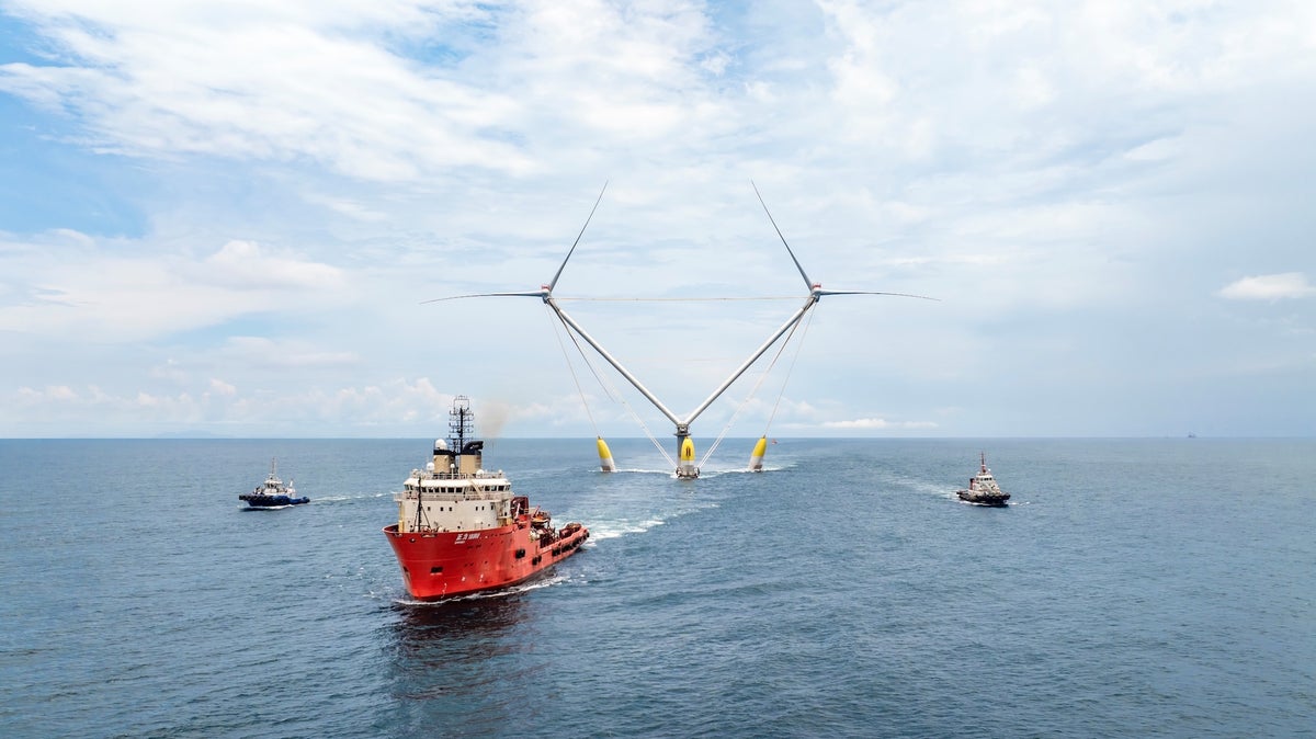 A red boat pulls an offshore wind turbine with two sets of blades anchored to a central platform over the ocean
