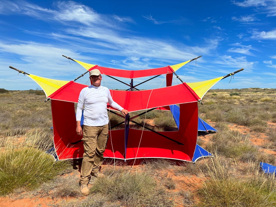 A person wearing a hat standing in front of a giant boxy kite. The kite has a red body, blue wings, and yellow fins. The landscape is a dry grassland and the sky is covered with thin clouds. 