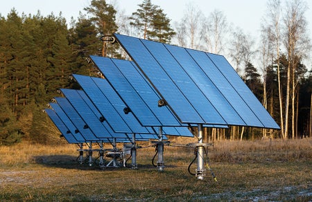 A row of siven vertical solar panel units mounted on poles in a grassy field dusted with snow.