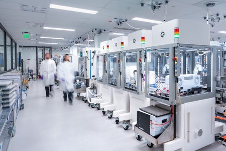 Technicians in white lab coats walk through a bright, modern robotic laboratory with rows of automated machines and enclosed workstations used for scientific processes.