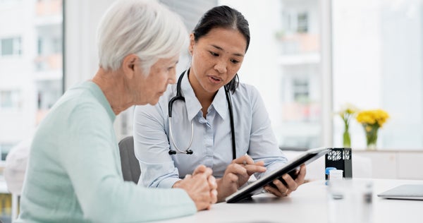 An elderly woman and a doctor with a stethoscope around her neck looking down at a tablet