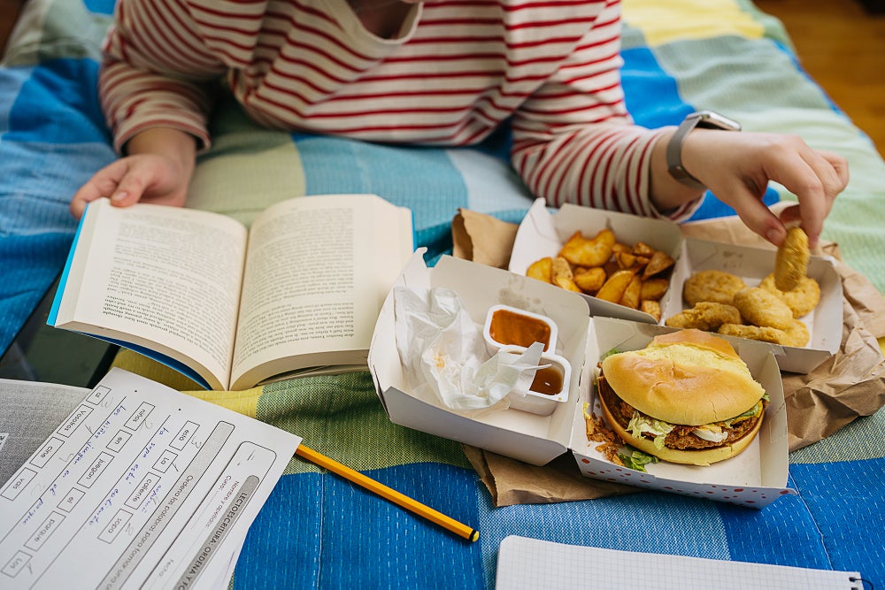 Mujer joven irreconocible acogedora en la cama, acostada boca abajo, apoyada en los codos, leyendo, estudiando y comiendo comida rápida en la cama