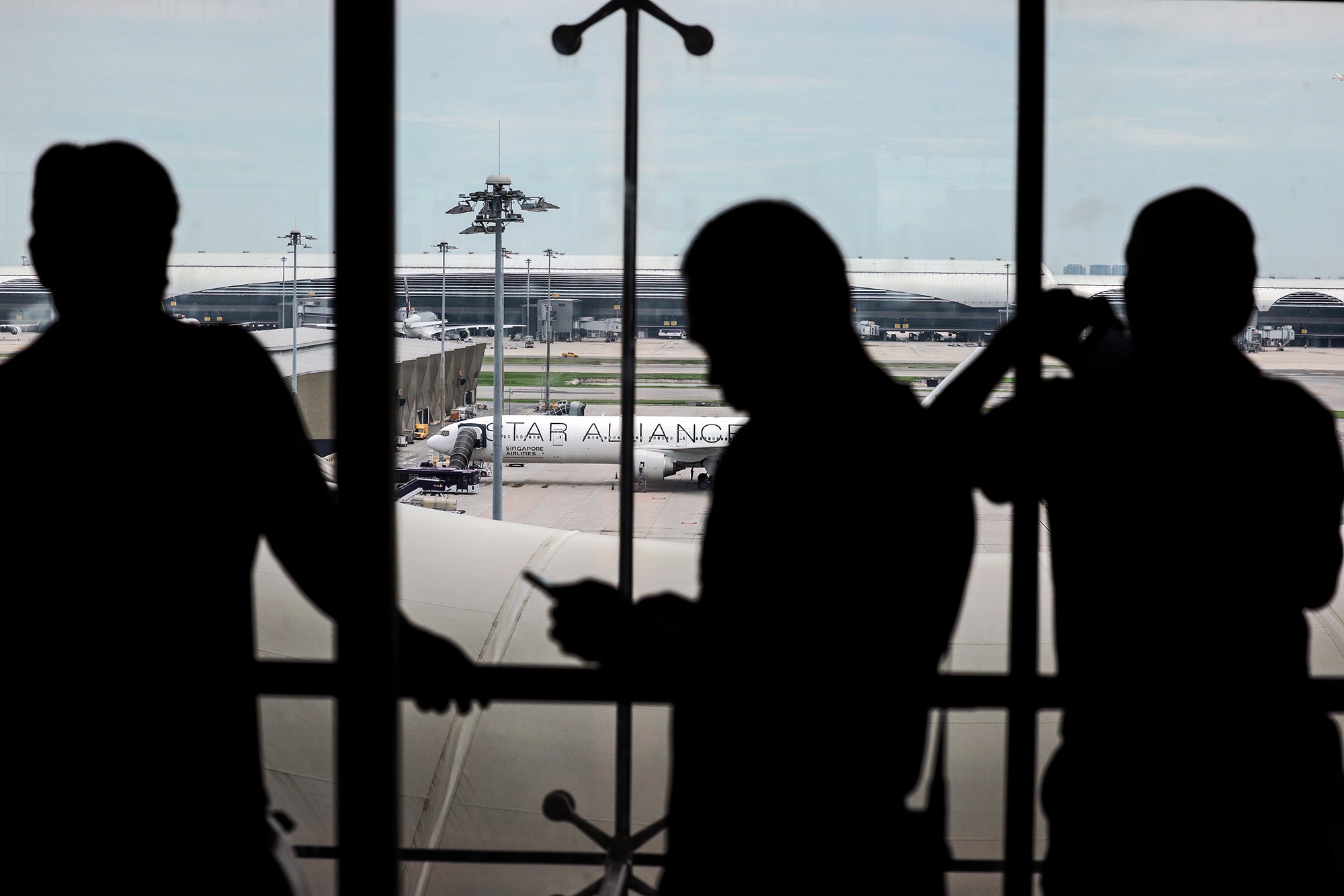 Photograph looking outwards from an airport window to a parked Signapore Airlines flight on the tarmac as passengers seen in silhouette stand in front of the window taking photos, looking at the plane, and using their phones