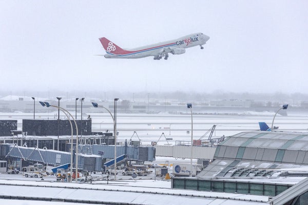 A plane takes off from a snowy runway with jet bridges and other airport infrastructure in the foreground