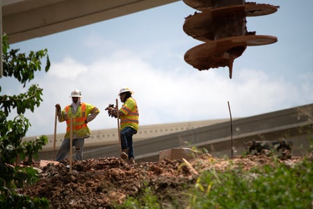 2 construction workers with shovels pausing under extreme heat with heavy equipment framing the photo.