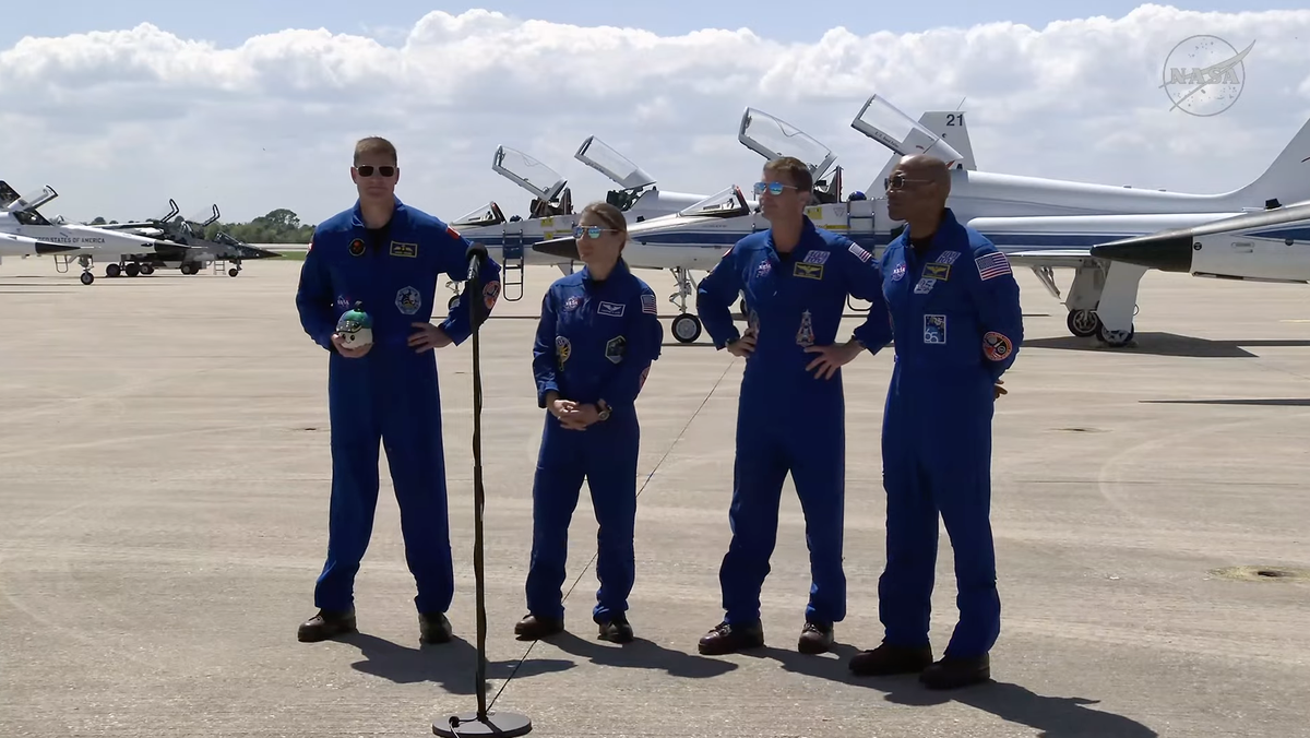Four people standing in blue flight suits arrayed behind a microphone.