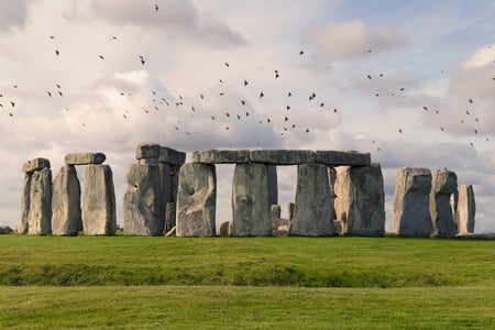 A flock of birds flying above Stonehenge on a partially cloudy day