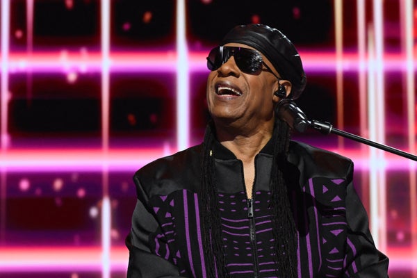 Stevie Wonder performs onstage against a pink geometric background