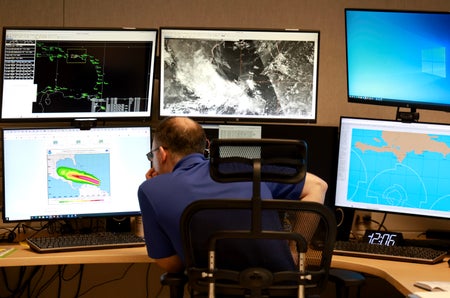 Hurricane specialist sitting at brown desk reviewing hurricane activity on 6 monitors